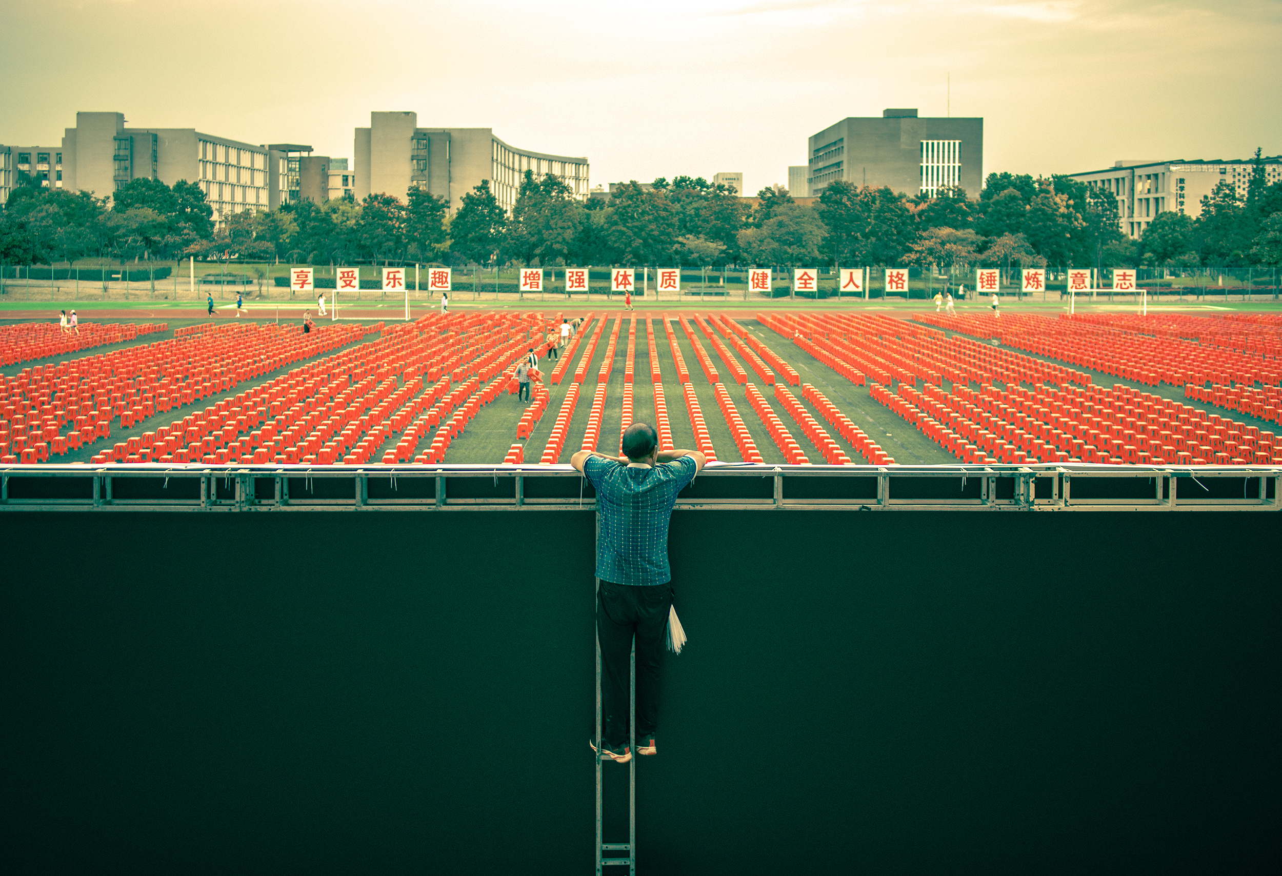 At a university stadium, a worker is laying a banner for the opening ceremony in two days. He leans over to watch the newly finished stadium. 