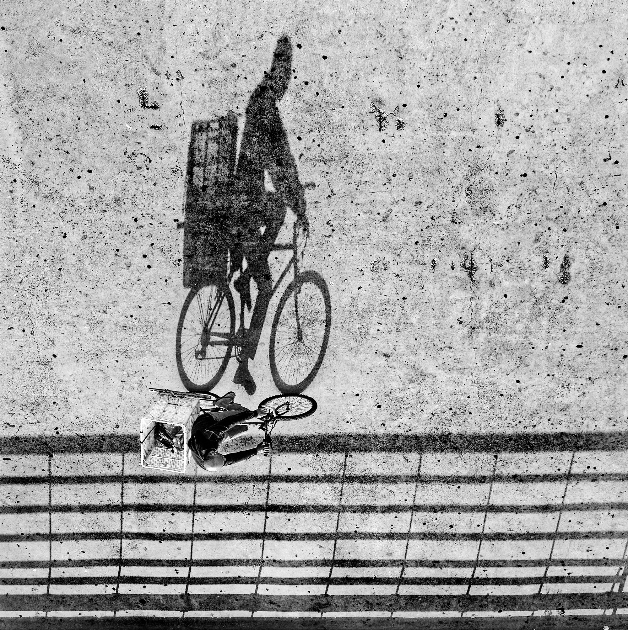 A man transports his work baskets on the back seat of his bicycle accompanied by his sharply defined shadow.