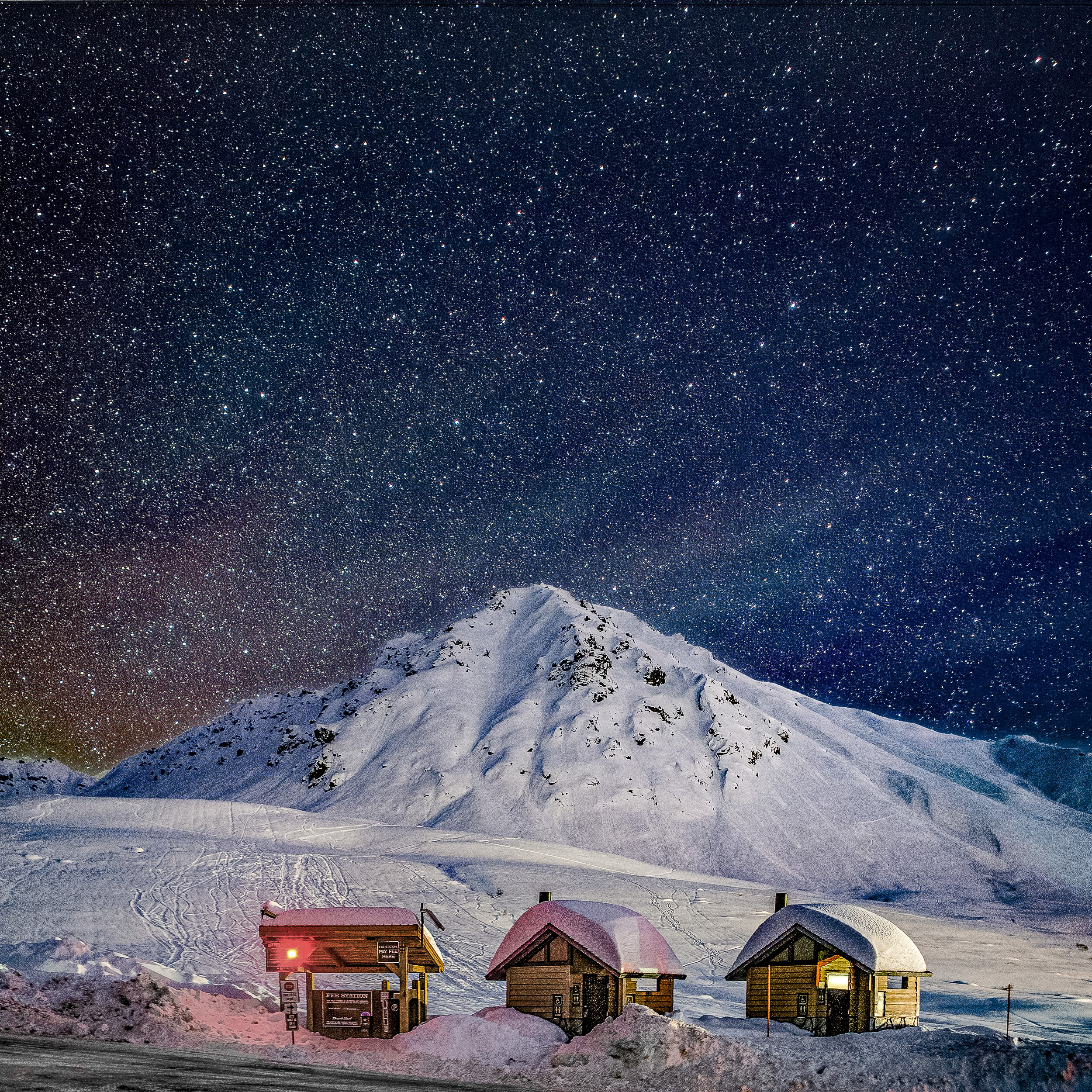 Starry skies and a hint of northern lights blaze over a snowy peak and three little chalets in Hatcher Pass, Alaska.