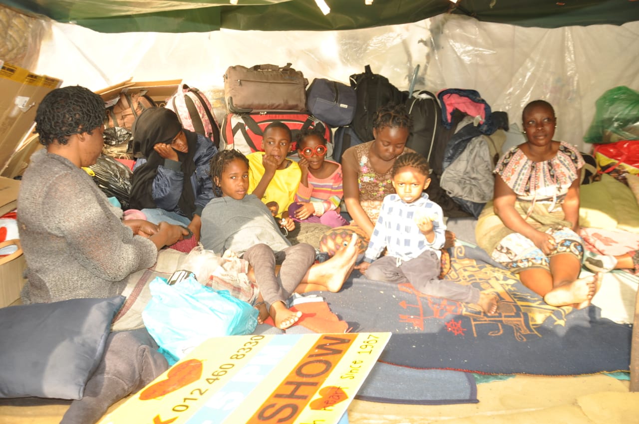 Refugee children and their mothers sit beneath a temporary shelter outside the UNHCR 