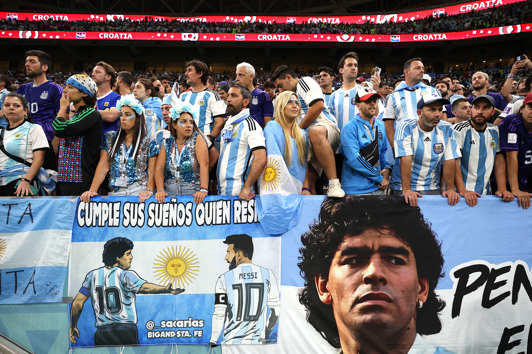 Fans of Argentina in front of a banner / flag showing the face of Diego Maradona and Lionel Messi 