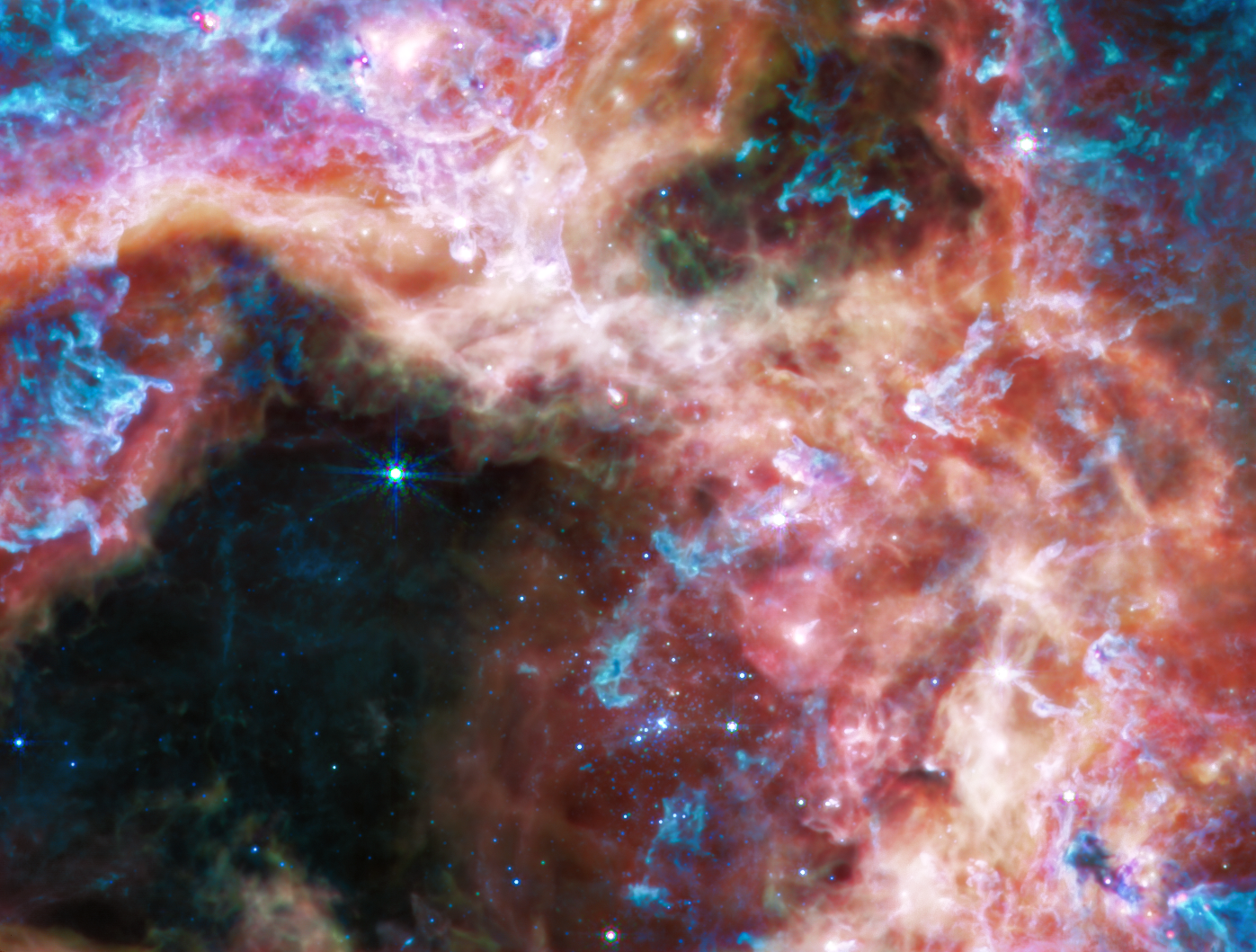 At the longer wavelengths of light captured by its Mid-Infrared Instrument (MIRI), Webb focuses on the area surrounding the central star cluster and unveils a very different view of the Tarantula Nebula. In this light, the young hot stars of the cluster fade in brilliance, and glowing gas and dust come forward. Abundant hydrocarbons light up the surfaces of the dust clouds, shown in blue and purple. Much of the nebula takes on a more ghostly, diffuse appearance because mid-infrared light is able to show more of what is happening deeper inside the clouds. Still-embedded protostars pop into view within their dusty cocoons, including a bright group at the very top edge of the image, left of center. Other areas appear dark, like in the lower-right corner of the image. This indicates the densest areas of dust in the nebula, that even mid-infrared wavelengths cannot penetrate. These could be the sites of future, or current, star formation. Image: NASA, ESA, CSA, STScI, Webb ERO Production Team