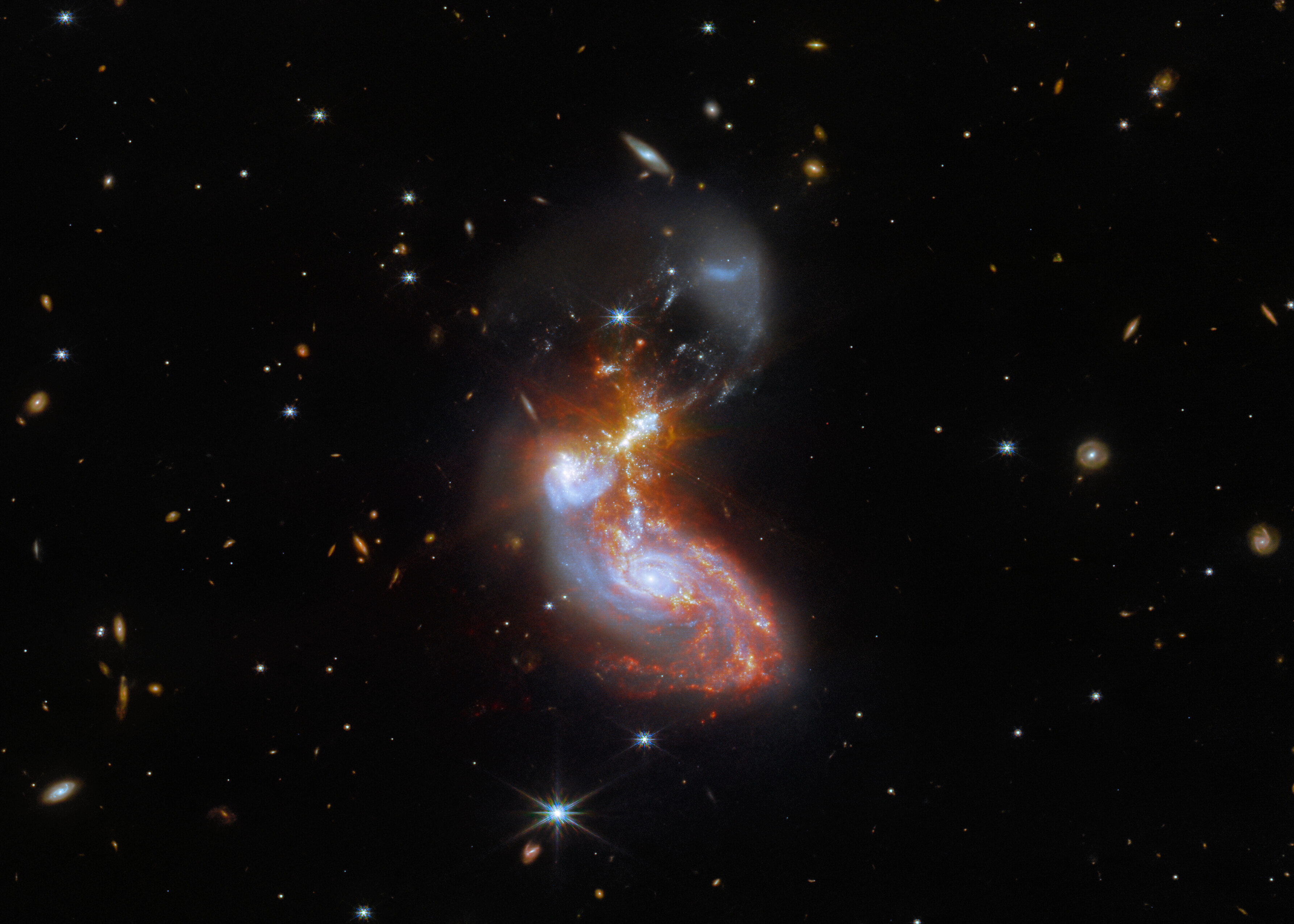A merging galaxy pair cavort in this image captured by the NASA/ESA/CSA James Webb Space Telescope. This pair of galaxies, known to astronomers as II ZW 96, is roughly 500 million light-years from Earth and lies in the constellation Delphinus, close to the celestial equator. As well as the wild swirl of the merging galaxies, a menagerie of background galaxies are dotted throughout the image. The two galaxies are in the process of merging and as a result have a chaotic, disturbed shape. The bright cores of the two galaxies are connected by bright tendrils of star-forming regions, and the spiral arms of the lower galaxy have been twisted out of shape by the gravitational perturbation of the galaxy merger. It is these star-forming regions that made II ZW 96 such a tempting target for Webb; the galaxy pair is particularly bright at infrared wavelengths thanks to the presence of the star formation. A galaxy merger lies in the centre of this image. The cores of the galaxies, coloured blue, are below-centre. They are surrounded by red star-forming regions which stretch up through and above the centre. Faint yellow diffraction spikes appear in the middle. The lower galaxy is a mostly regular spiral shape, while the upper galaxy has been distorted heavily. The background is black, and covered with many tiny galaxies throughout the scene