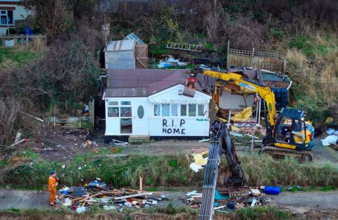 In this aerial view a beach chalet adorned with 'RIP HOME' is demolished by workers before it falls down the sea cliff on The Marrams at Hemsby Beach on December 11, 2023 in Hemsby, England. The collapse of a private access road in November, prompted by high tides and winds, led Great Yarmouth Borough Council to declare the rapidly eroding homes on The Marrams 'not structurally sound and unsafe.' This demolition coincides with the 10th anniversary of five village houses lost to the sea in 2013 when storms undermined foundations by stripping away significant sand. (Photo by Christopher Furlong/Getty Images)