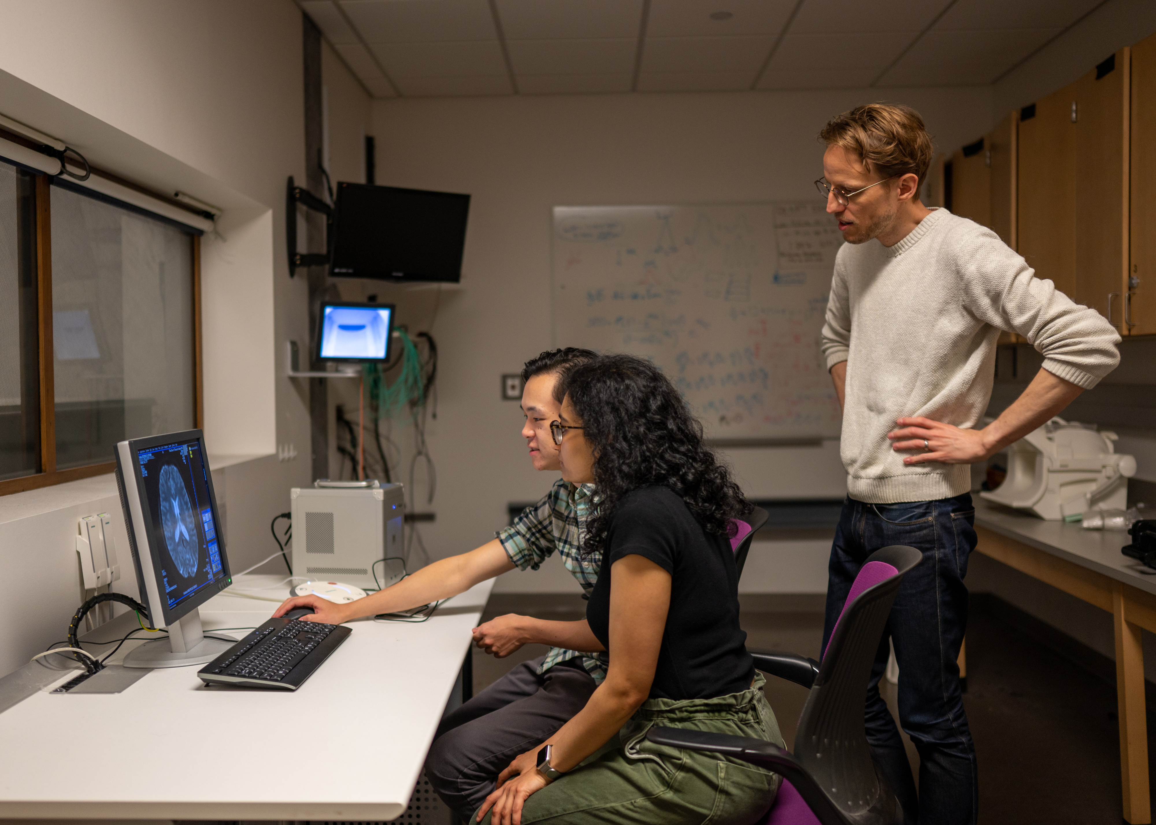 Alex Huth (standing), discusses the semantic decoder project with Jerry Tang (left) and Shailee Jain (right) in the Biomedical Imaging Center at the University of Texas at Austin. The researchers trained their semantic decoder on dozens of hours of brain activity data from participants, collected in an FMRI scanner. Image: Nolan Zunk/University of Texas at Austin.